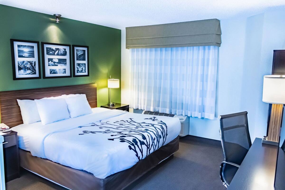 Quarto de hotel com cama, escrivaninha, cadeira, abajur e janela com cortinas.