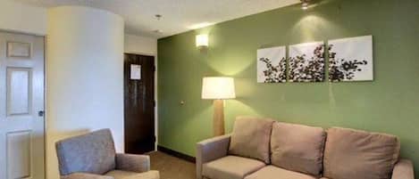 A room with a beige sofa, a small armchair, a coffee table, a lamp, and a wall with a floral artwork.