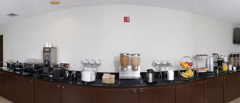A breakfast bar with coffee machines, a fruit basket, and various food containers.