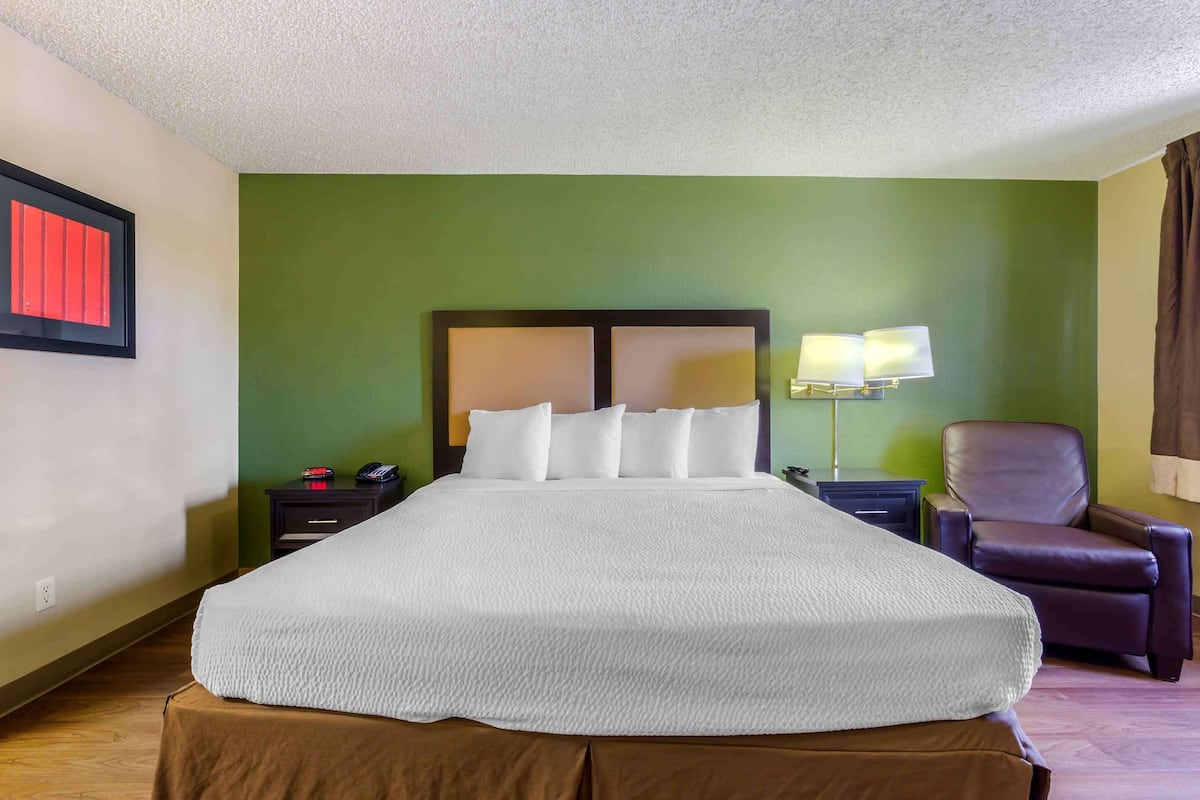 A hotel room with a large bed, a nightstand, a lamp, a chair, and a red-framed window.