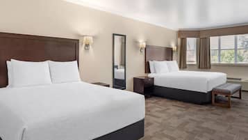 A hotel room with two beds, a large mirror, bedside lamps, a bench, and a window with blinds.