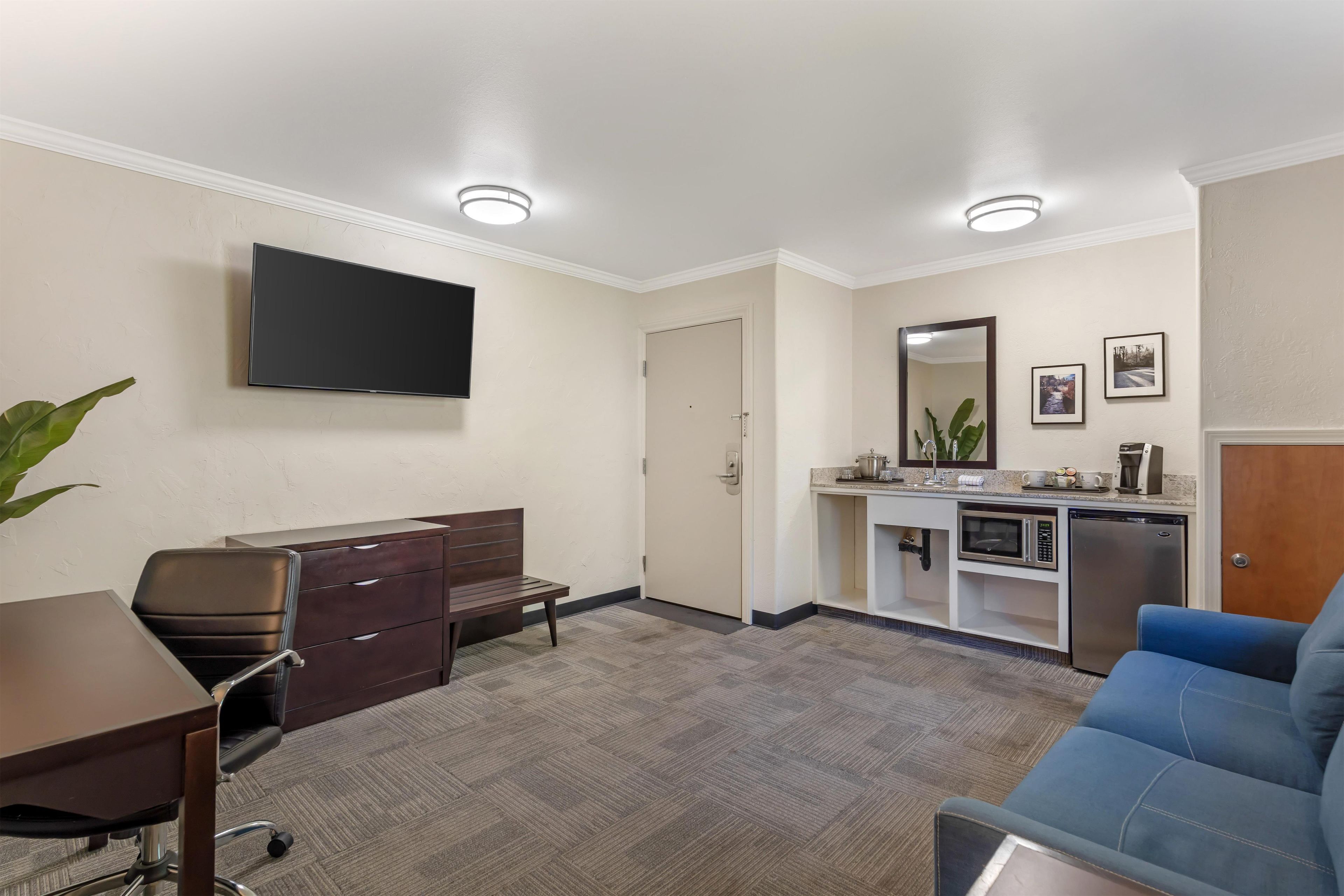 Habitación de hotel con televisor de pantalla plana, escritorio, silla, sofá, kitchenette con microondas y refrigerador, y una puerta que da a otra habitación.