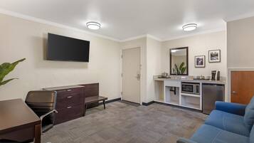 Habitación de hotel con televisor de pantalla plana, escritorio, silla, sofá, kitchenette con microondas y refrigerador, y una puerta que da a otra habitación.