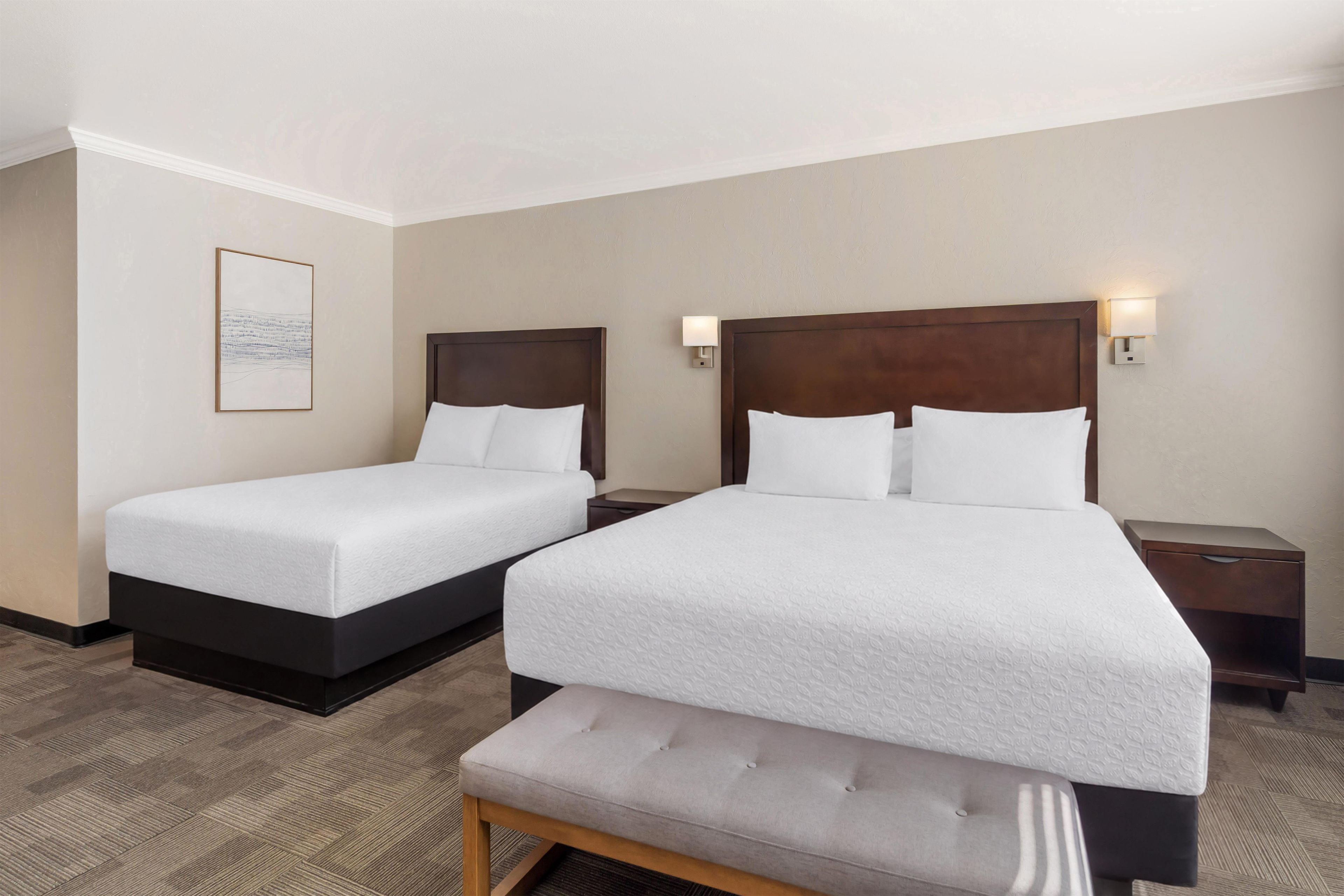 A hotel room with two beds, a bench, and a nightstand with a lamp.