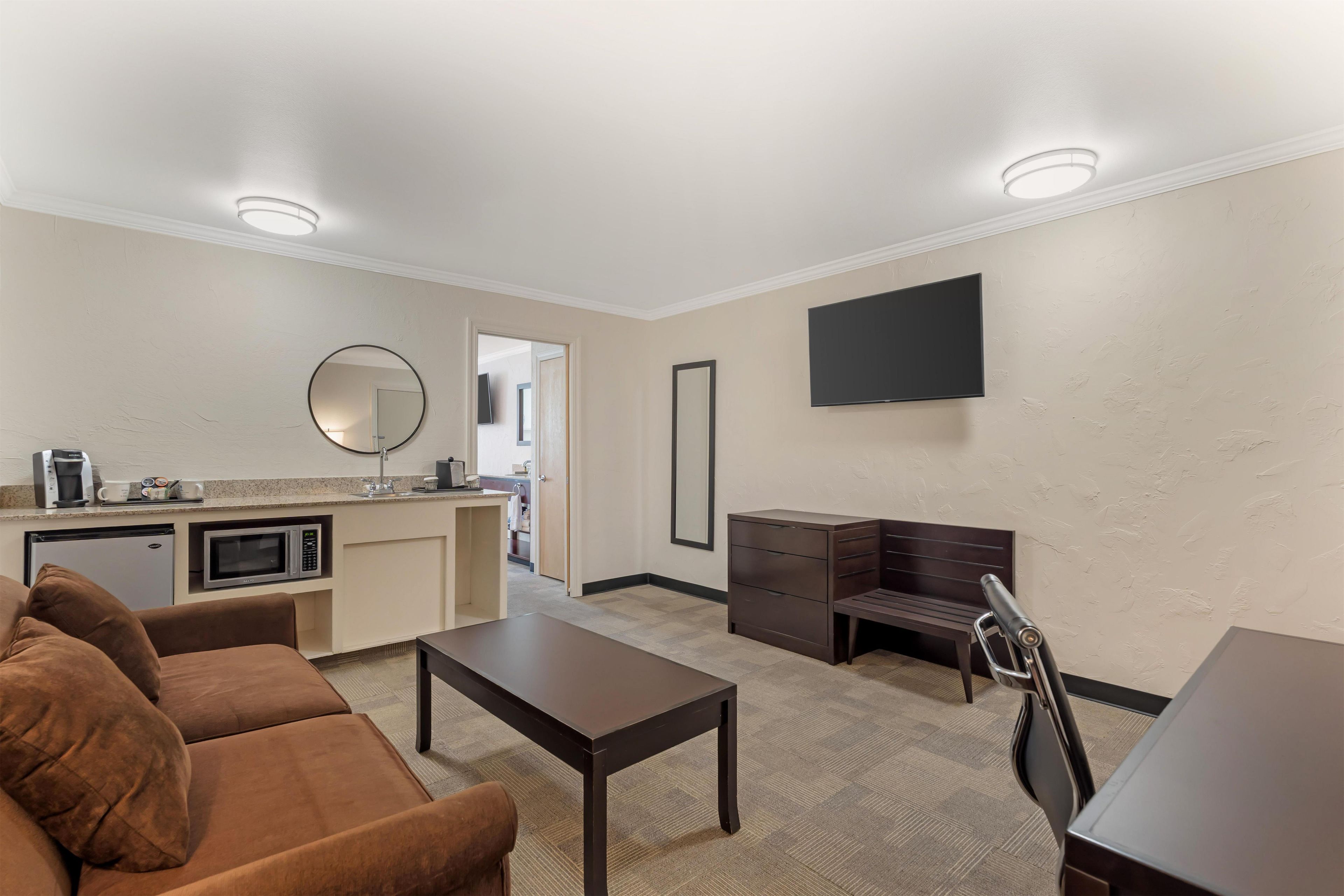 A modern living room with a brown sofa, a coffee machine, a microwave, a flat-screen TV, and a desk with drawers.