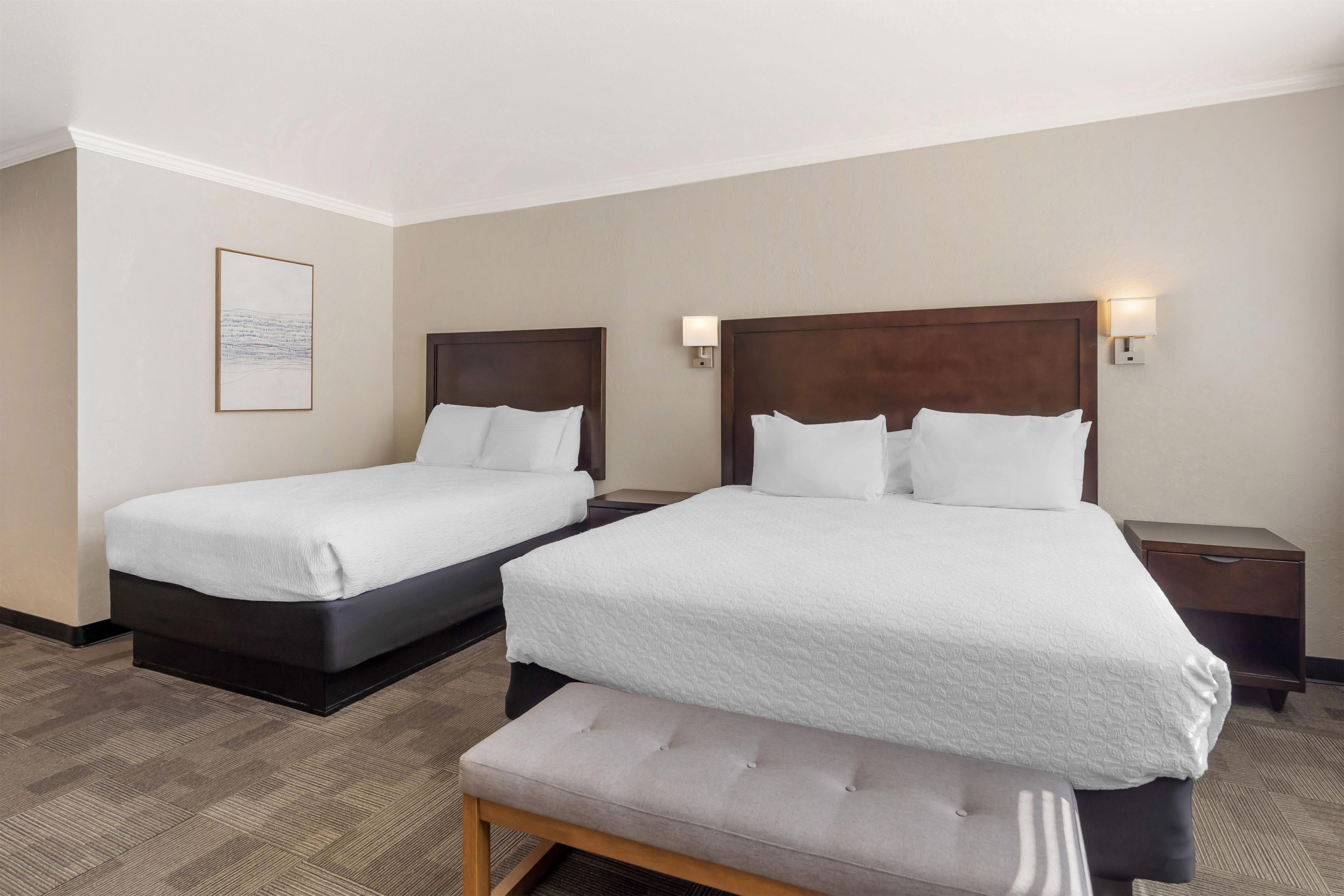 A hotel room with two beds, a bench, and a framed picture on the wall.
