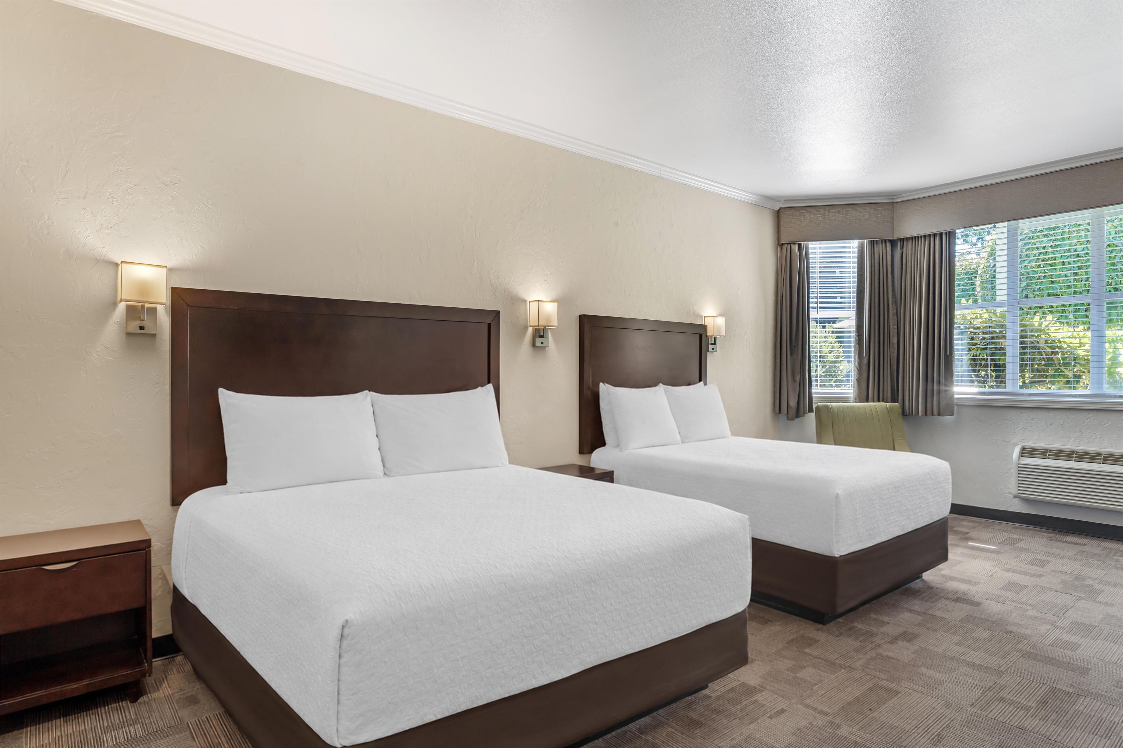 A hotel room with two beds, a nightstand, a chair, a window with curtains, and a wall-mounted heater.