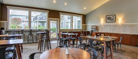 A spacious dining area with multiple tables and chairs, large windows, and a kitchen area with wooden cabinets.