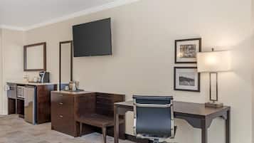 A hotel room with a desk, chair, flat-screen TV, and a small kitchenette.