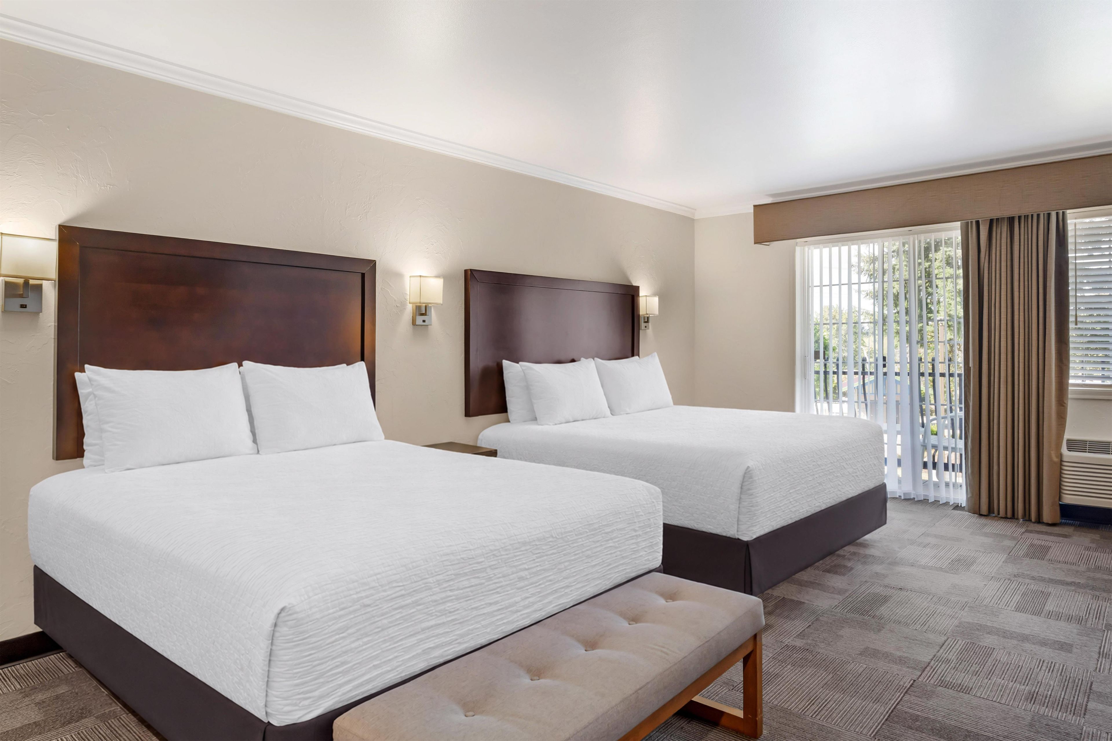 A hotel room with two beds, a bench, and a view of the outdoors.