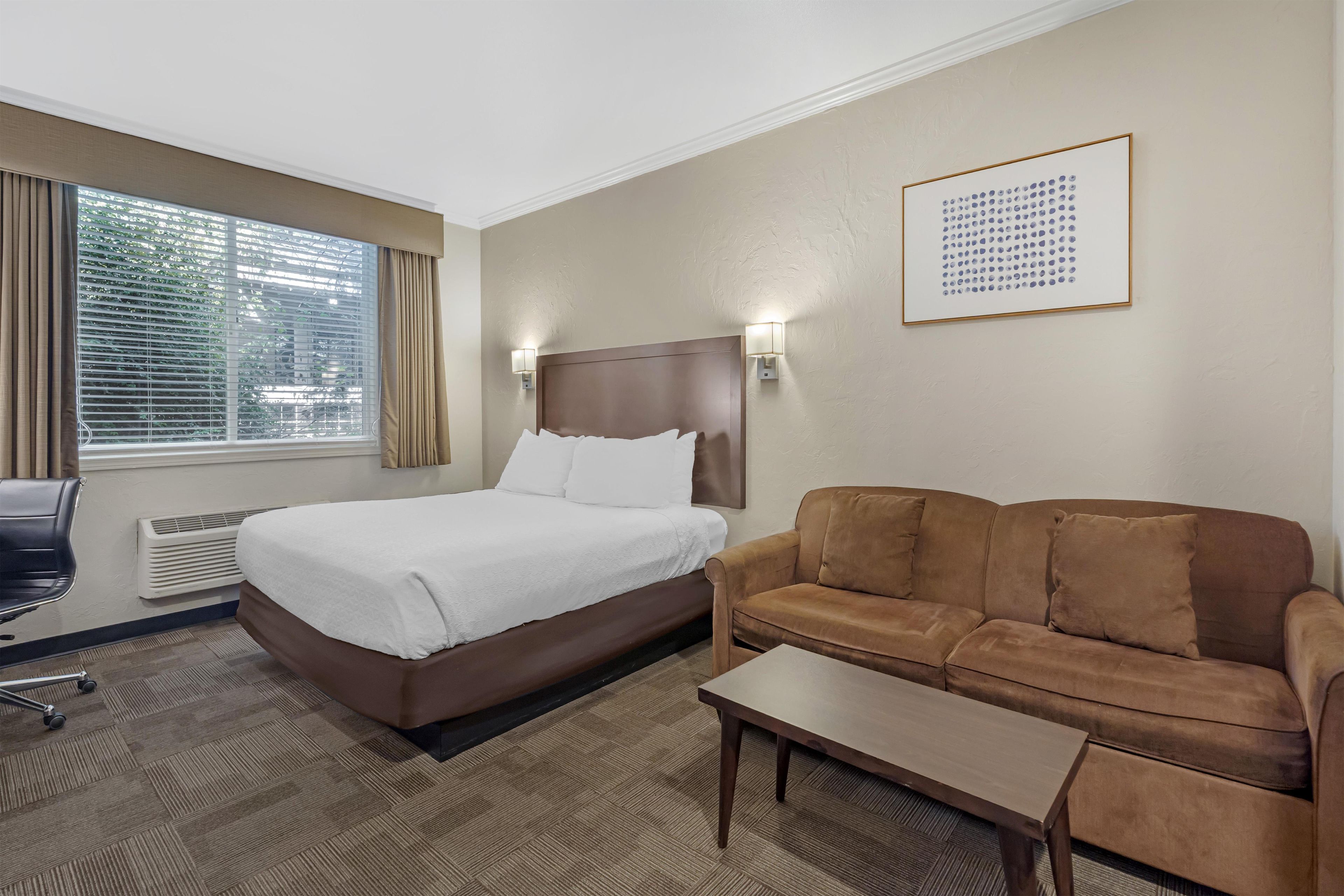 Habitación de hotel con cama, sofá marrón, una mesita, una silla y ventana con persianas.