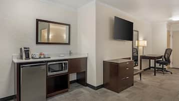 A compact hotel room with a kitchenette, a flat-screen TV, a desk with a chair, and a mirror.