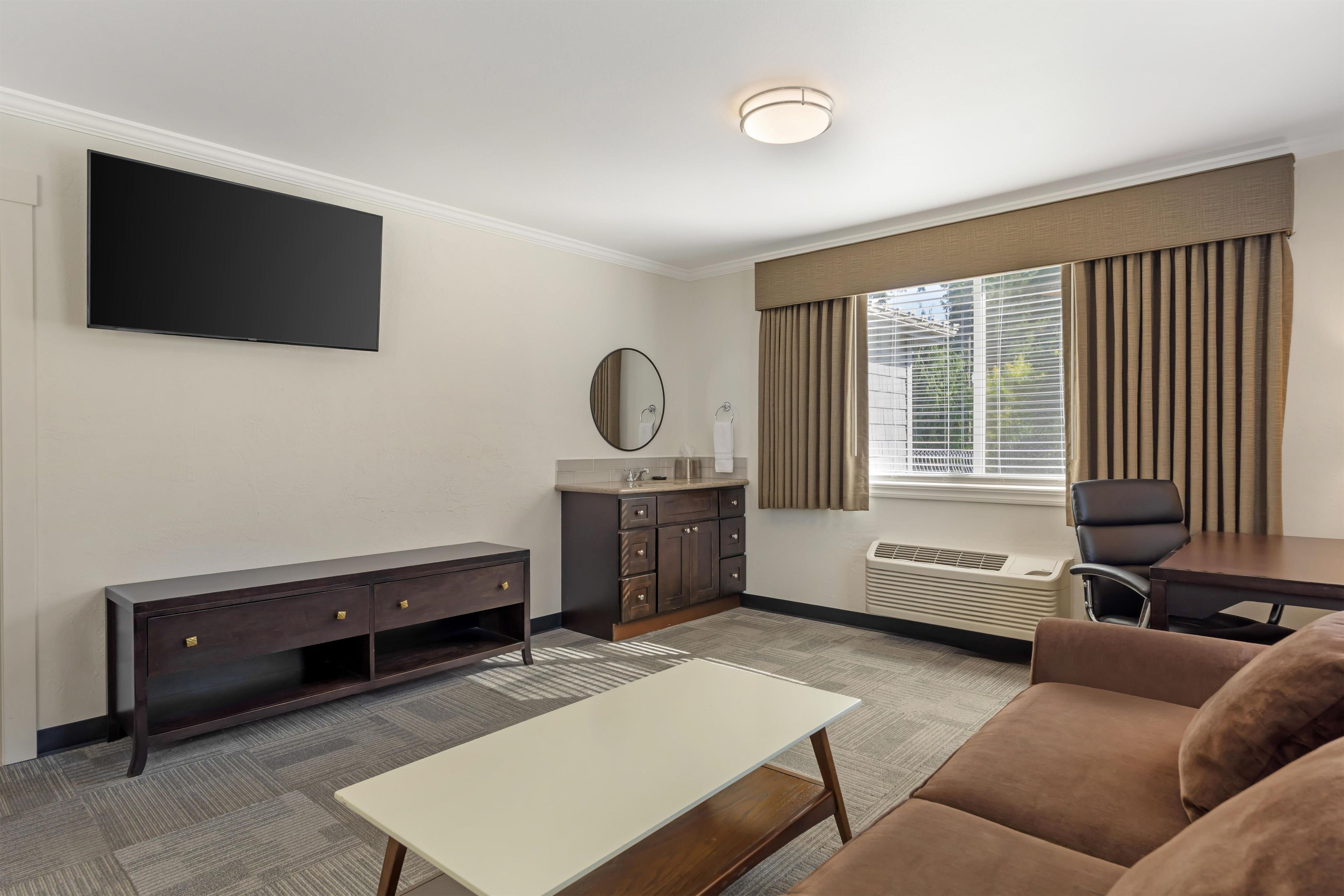 Ein Hotelzimmer mit Flachbildfernseher, einer Sitzecke, einem Schreibtisch, einem Spiegel, einem Schminktisch und einem Fenster mit Jalousien.
