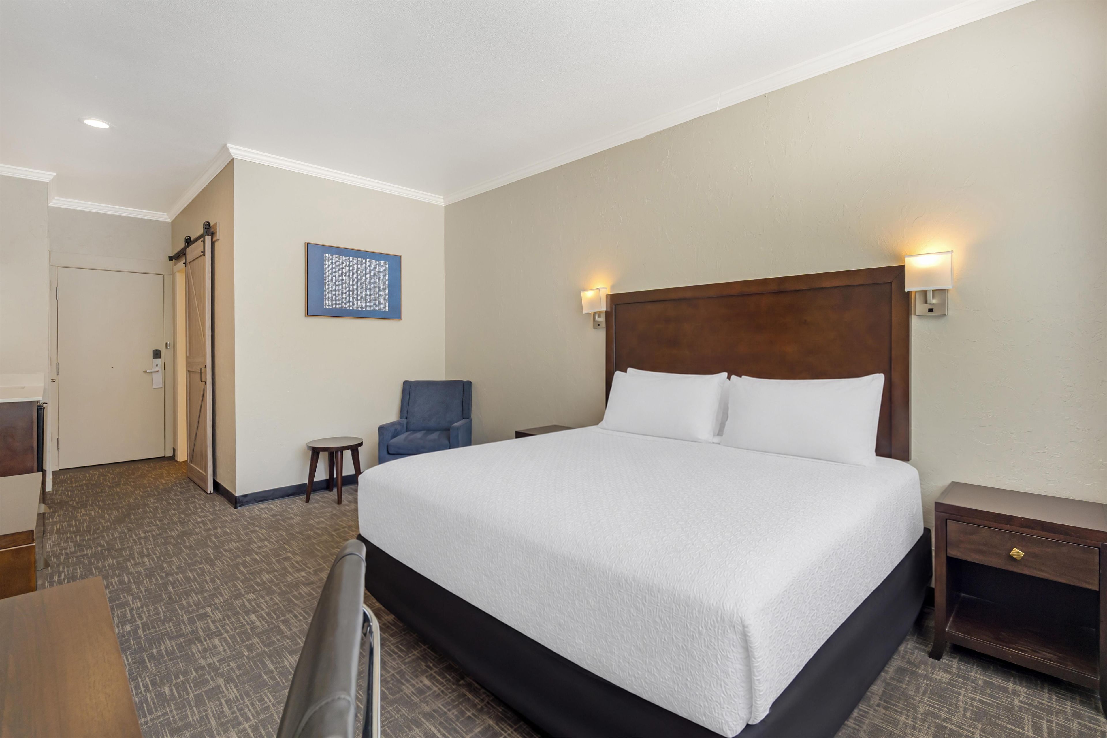 A hotel room with a large bed, a chair, a desk, and a wall-mounted light fixture.