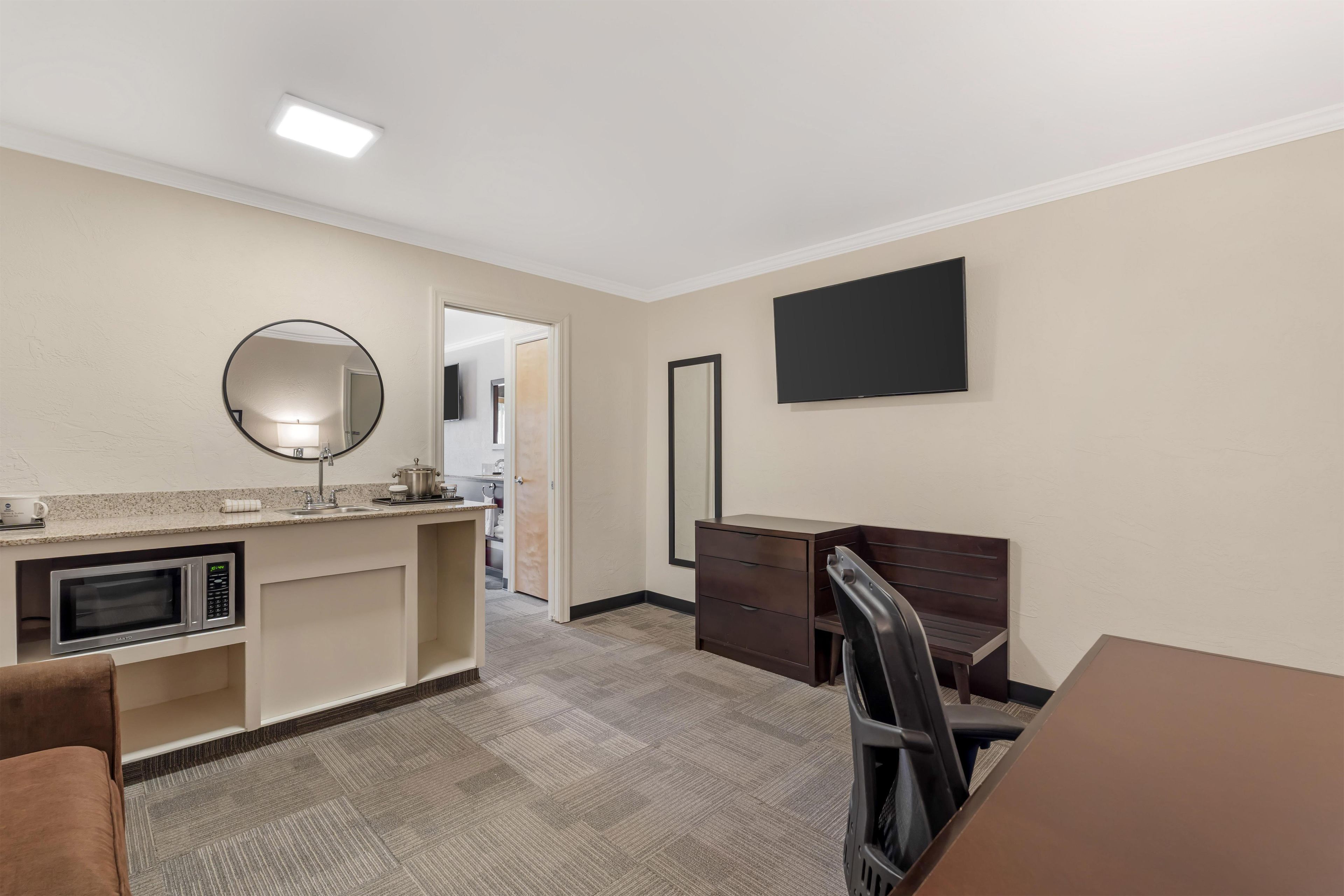 A hotel room with a kitchenette, a flat-screen TV, a mirror, and a desk with a chair.