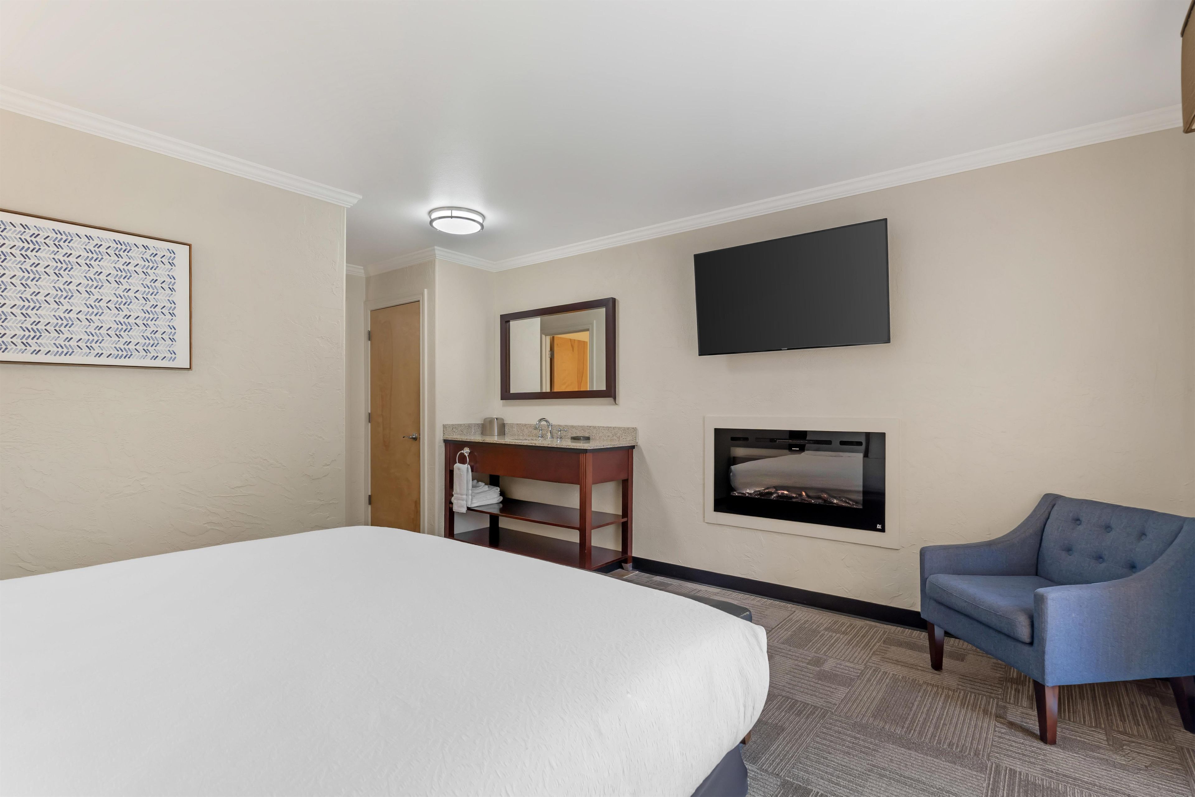 A hotel room with a bed, a desk with a mirror, a flat-screen TV, and a fireplace.