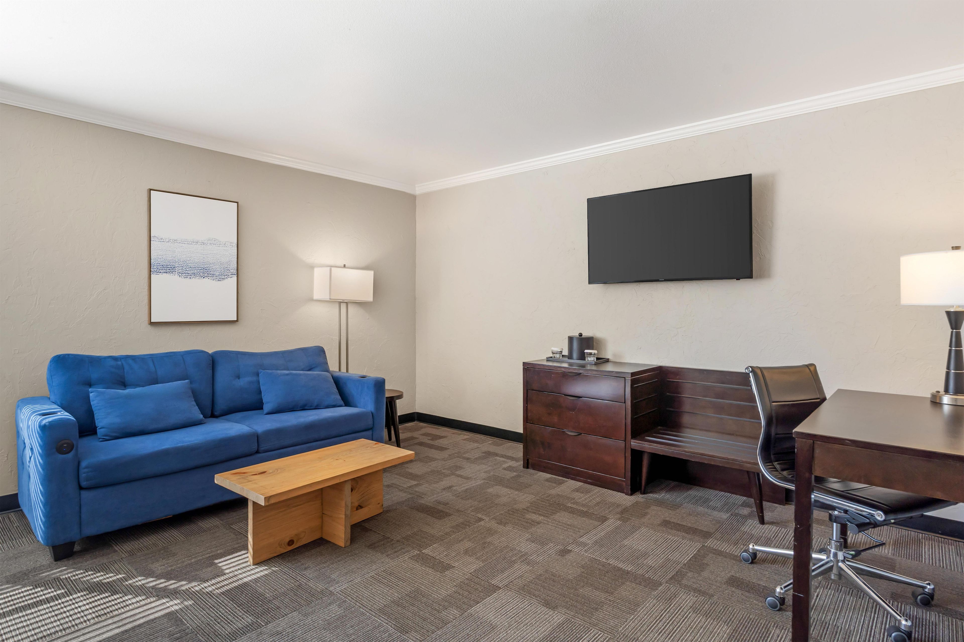 A hotel room with a blue sofa, a wooden coffee table, a desk with a chair, a flat-screen TV, and a lamp.