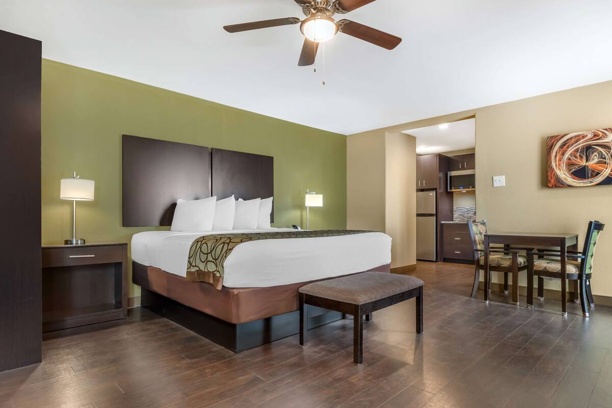 A hotel room with a large bed, bedside tables, a ceiling fan, a dining area with chairs, and a kitchen in the background.