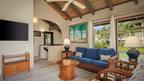 A living room with a blue sofa, wicker furniture, a glass coffee table, a flat-screen TV, and a painting of palm trees. - Aston at Poipu Kai (Koloa)