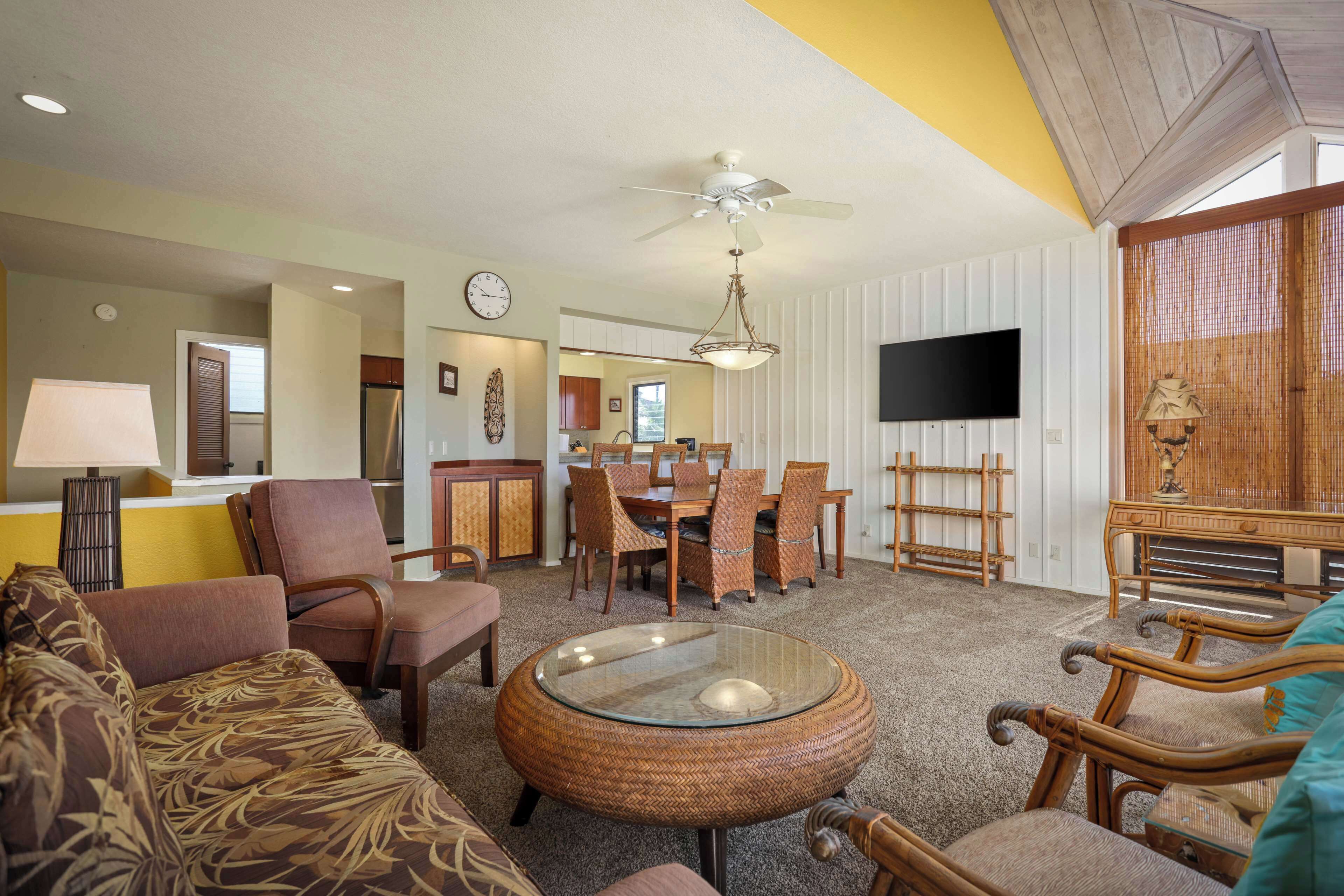 A spacious living room with a round glass coffee table, comfortable seating, a dining area with a table and chairs, a ceiling fan, and a flat-screen TV mounted on the wall.