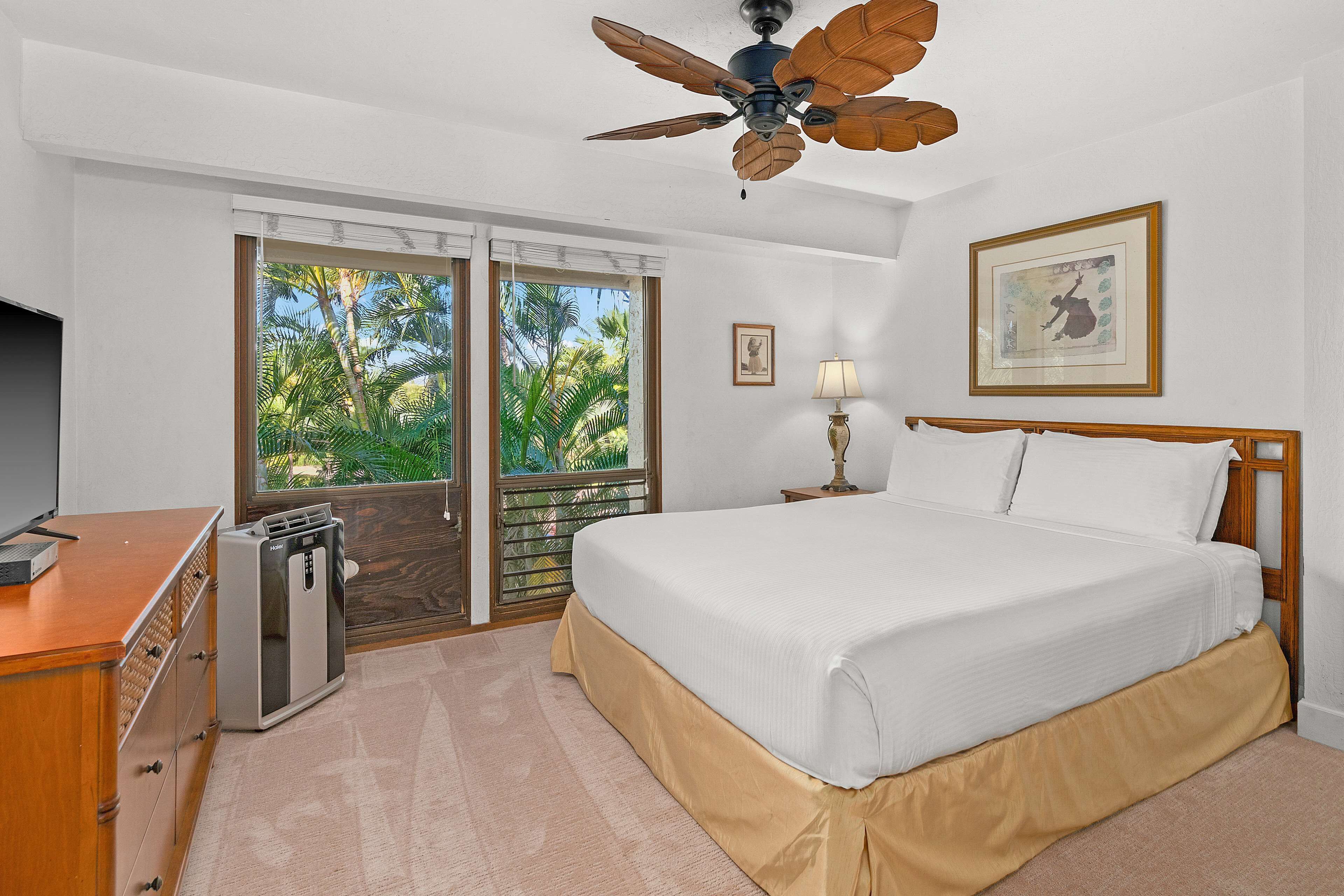 A hotel room with a bed, a desk, a ceiling fan, a window with a view of palm trees, and a framed picture on the wall.
