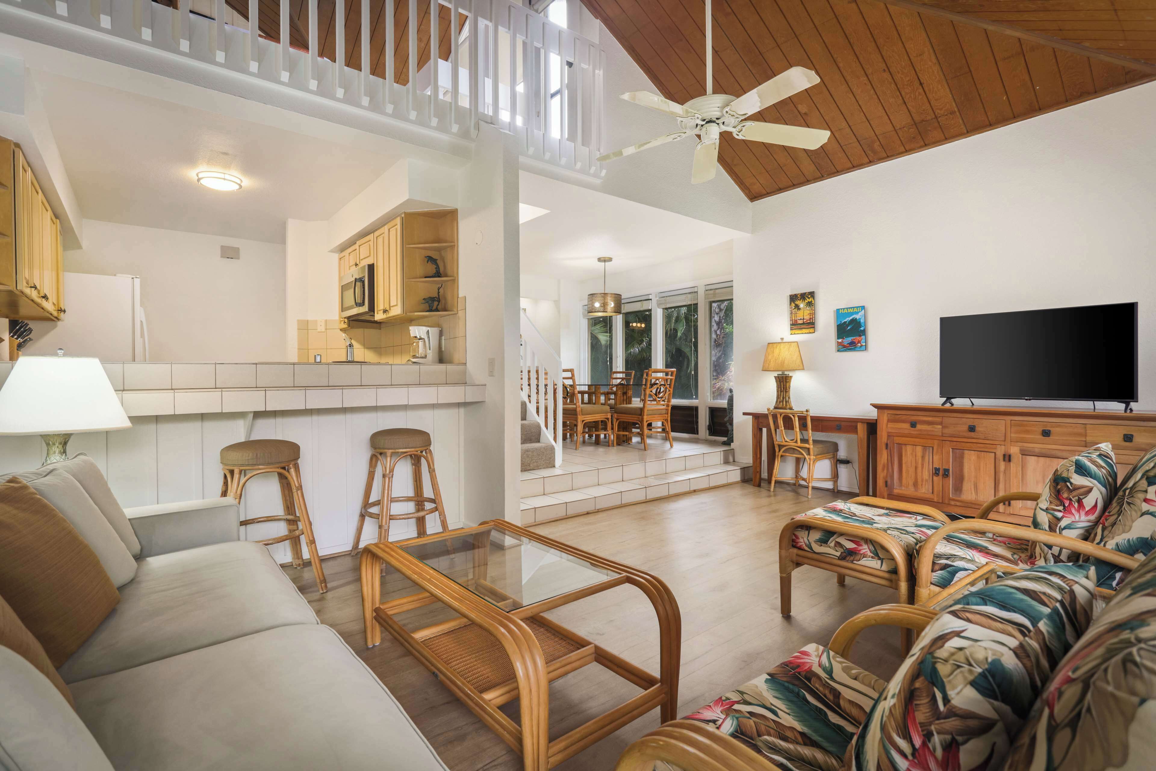 A spacious living area with a sofa, a glass coffee table, and a TV. There is a kitchen area with wooden cabinets and a microwave.