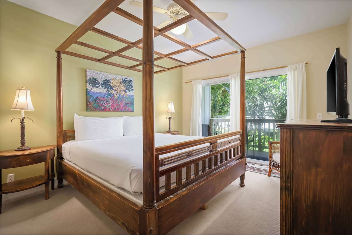 A four-poster bed with a wooden frame, two bedside tables with lamps, a painting on the wall, a television on a wooden cabinet, and a balcony with a view of greenery.