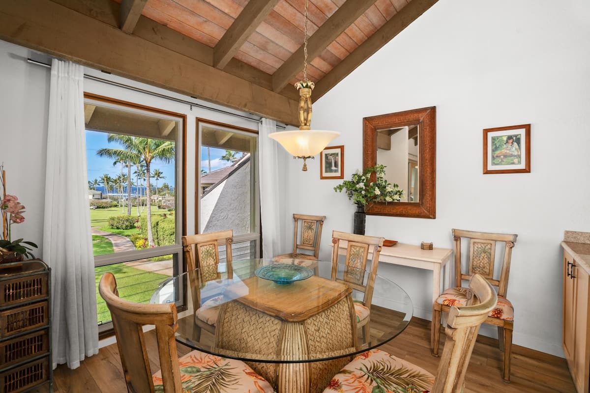A dining area with a glass table, wooden chairs, a hanging light fixture, and a view of a garden through large windows.