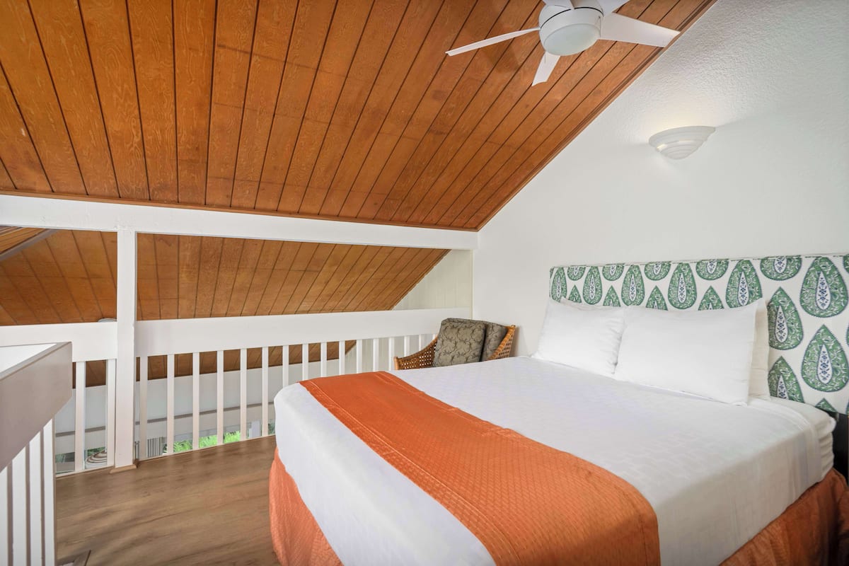 A bedroom with a wooden ceiling, a bed with white and orange bedding, a patterned headboard, and a ceiling fan.