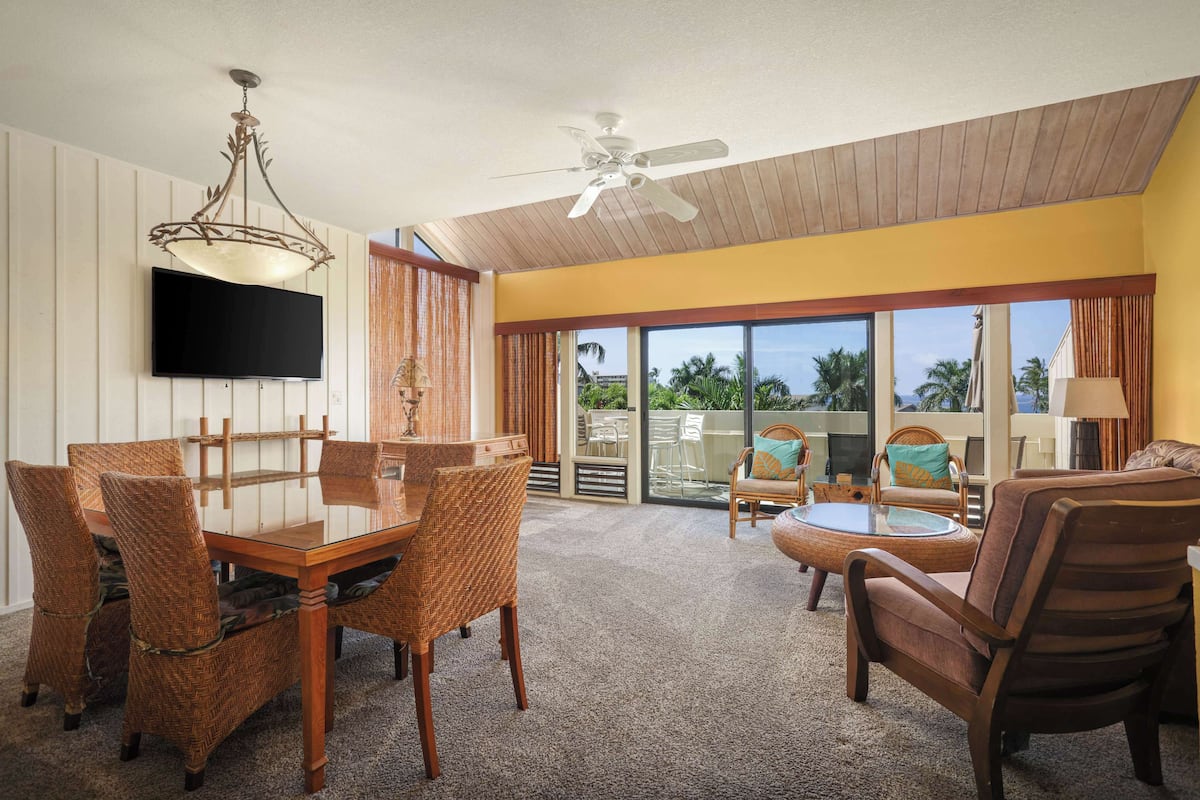 A spacious living area with a dining table, wicker chairs, a ceiling fan, and a large window offering a view of palm trees.