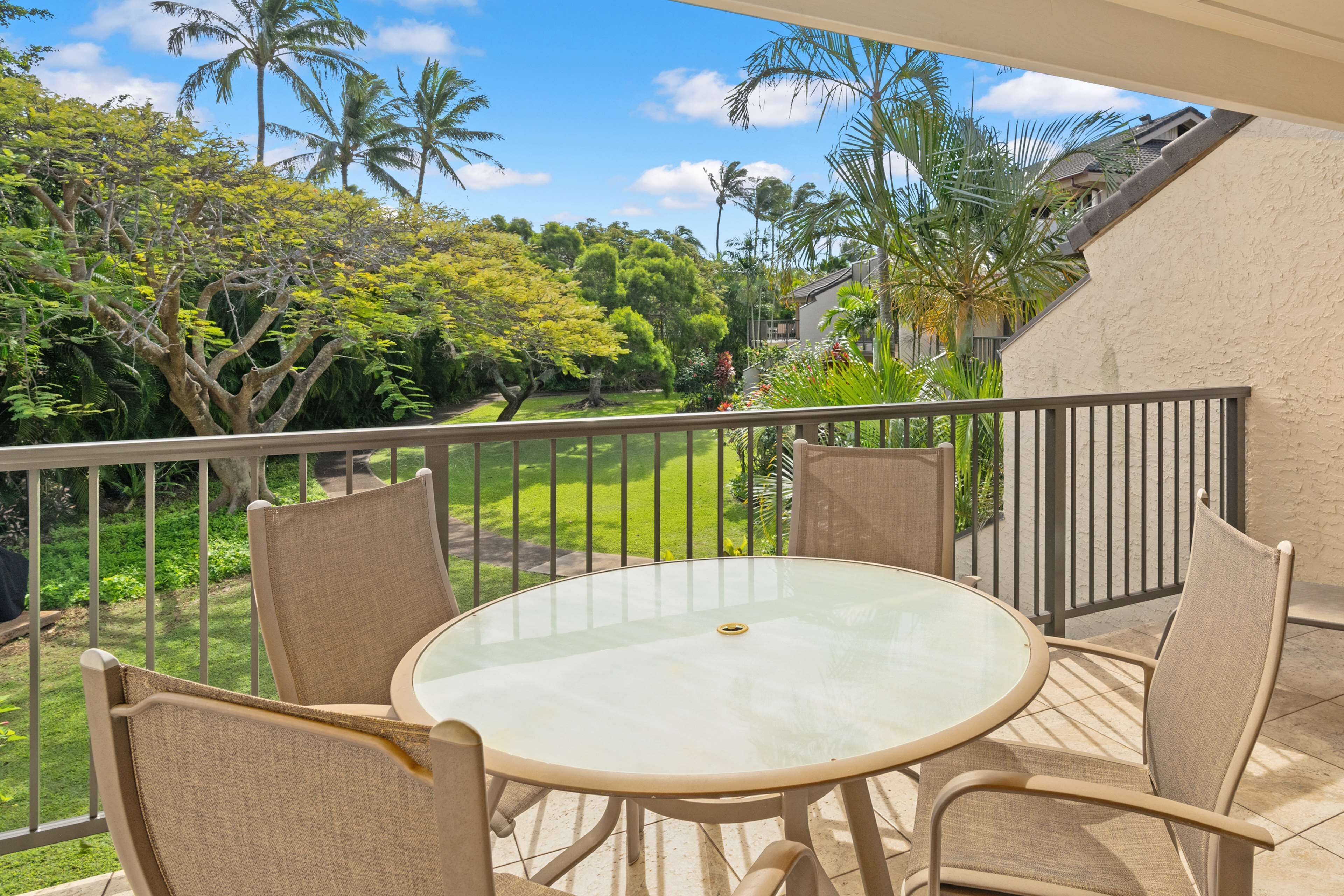 Un balcon doté d’une table ronde en verre et de quatre chaises, offrant une vue sur un jardin luxuriant, avec des palmiers et un ciel d’un bleu éclatant.