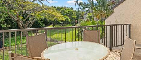 Un balcon doté d’une table ronde en verre et de quatre chaises, offrant une vue sur un jardin luxuriant, avec des palmiers et un ciel d’un bleu éclatant.