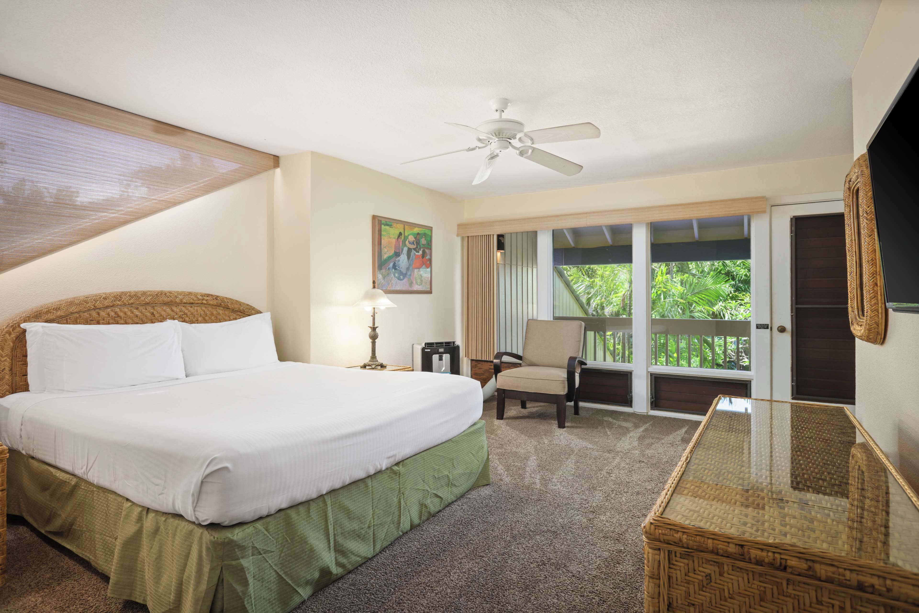 A hotel room with a large bed, a chair, a table, a ceiling fan, and a balcony with a view of greenery.