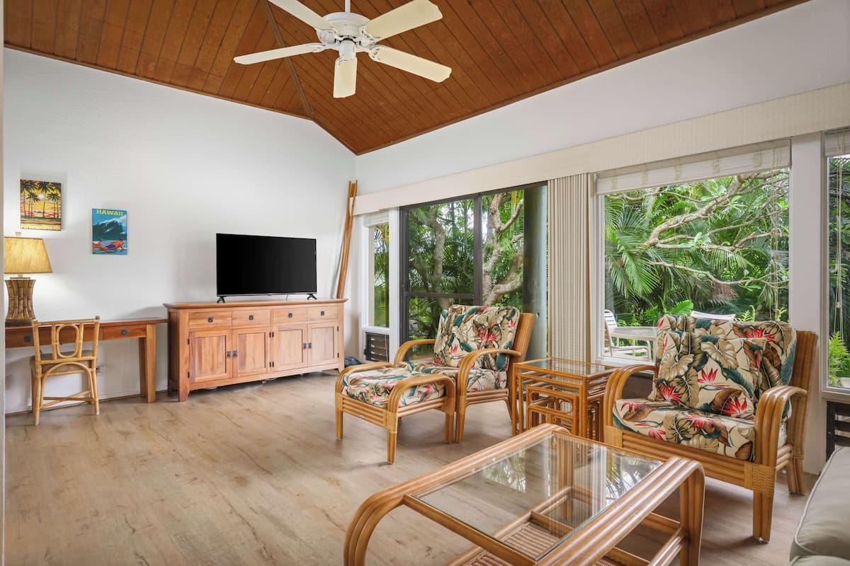 A spacious living room with a wooden ceiling, a television, wicker furniture, and large windows offering a view of tropical foliage.