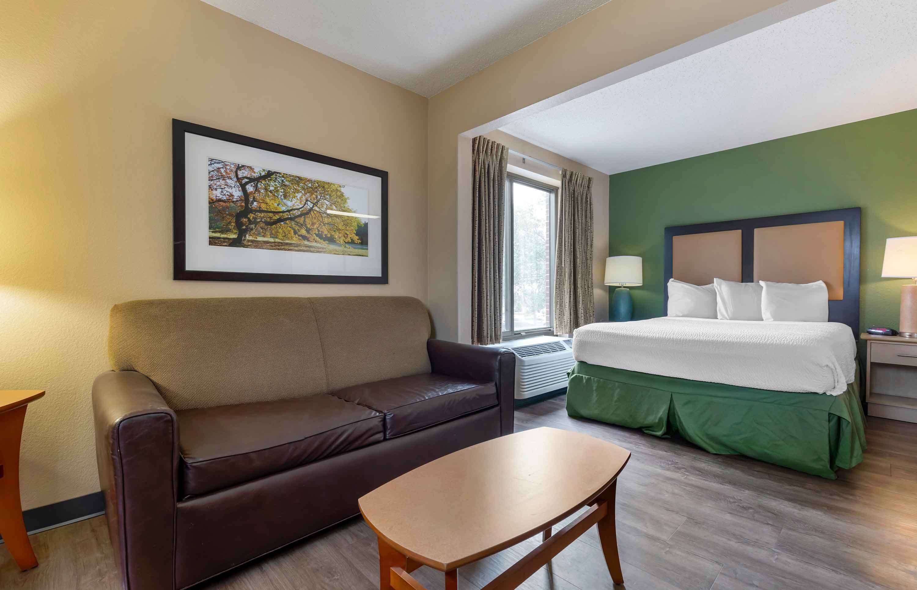 A hotel room with a bed, a brown leather sofa, a wooden table, a framed picture on the wall, and a window with curtains.