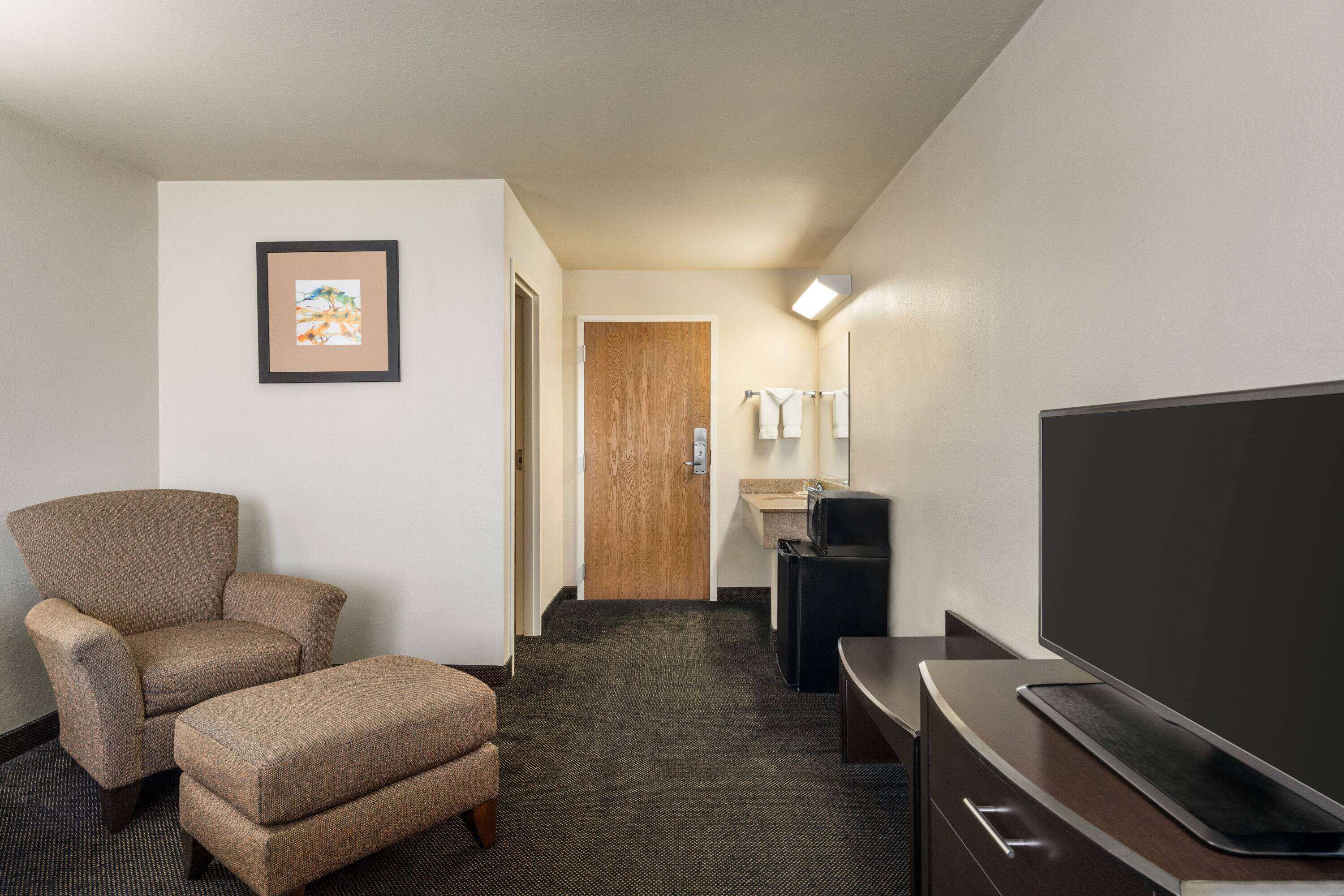 A hotel room with a large flat-screen TV, a sofa, a small ottoman, a framed picture on the wall, a wooden door, and a small sink area.