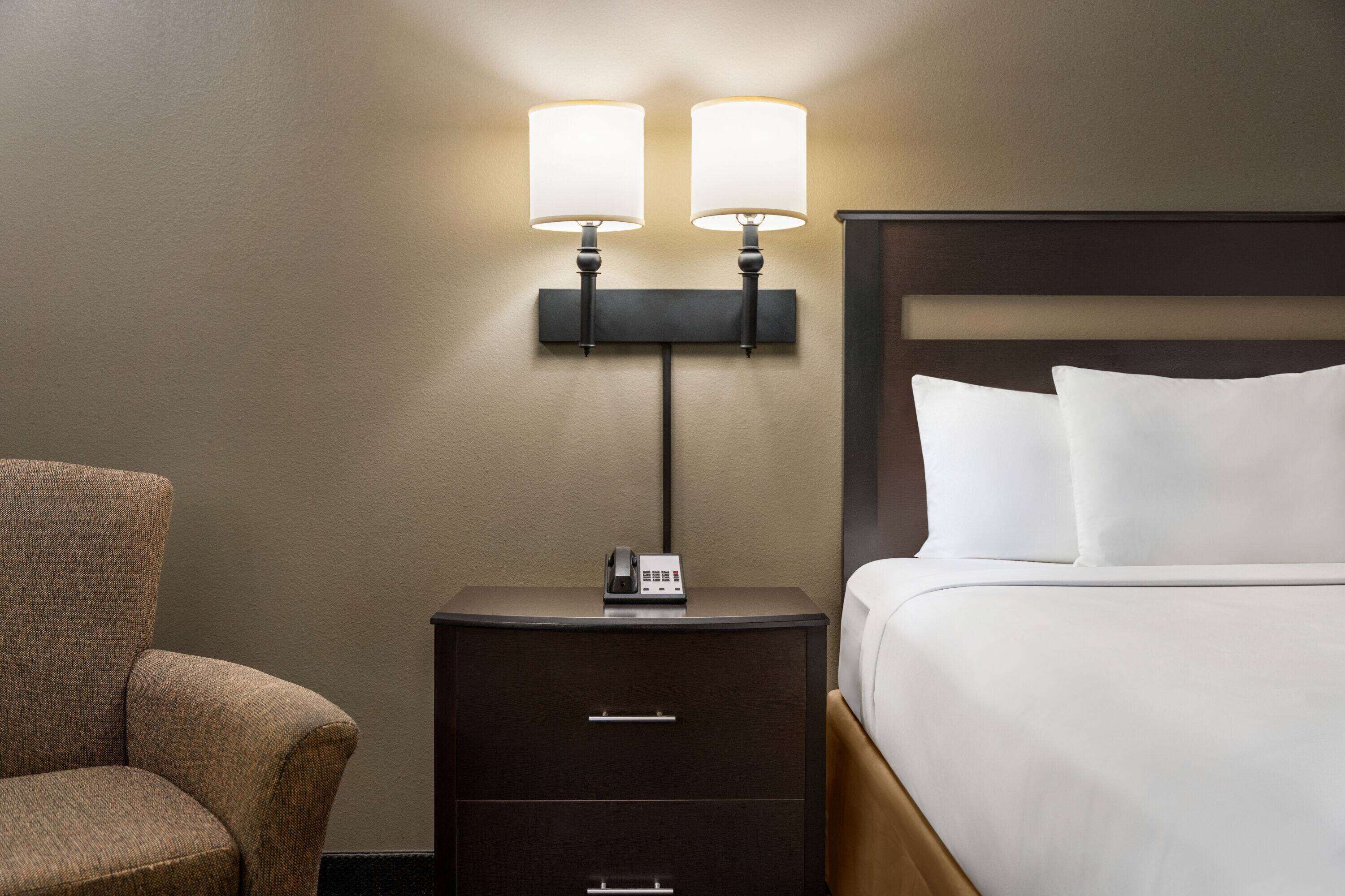 A hotel room with a bed, a nightstand with a phone, and a wall-mounted lamp.