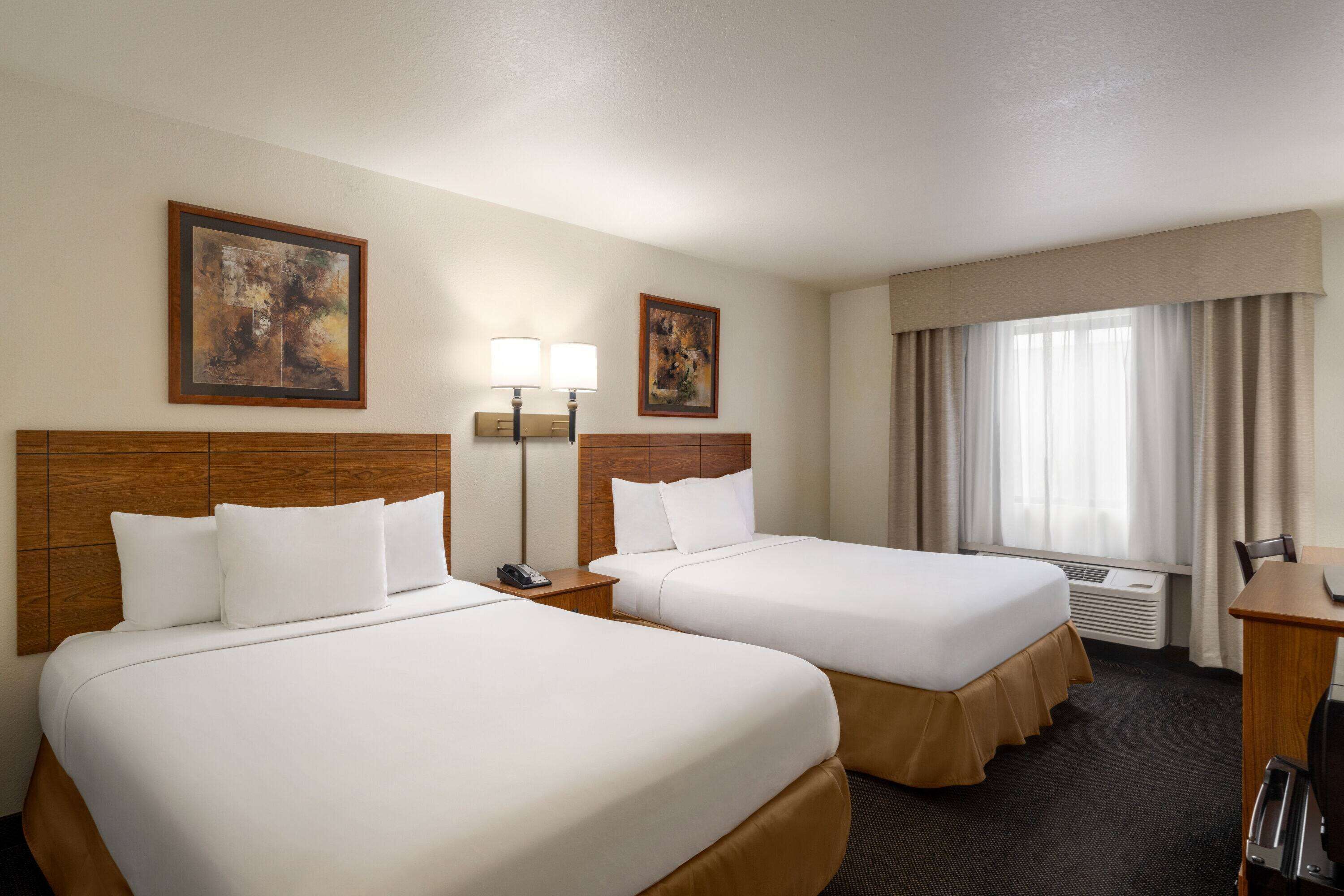 A hotel room with two beds, a desk, a chair, a lamp, and a window with curtains.