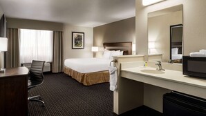 Habitación de hotel con cama, escritorio, silla, espejo grande, lavamanos y un cesto de basura.