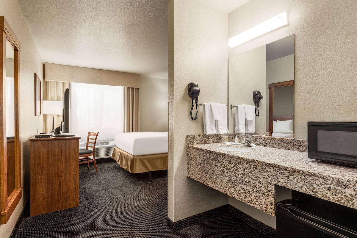 A hotel room with a bed, a desk, a chair, a vanity with a mirror, and a TV.