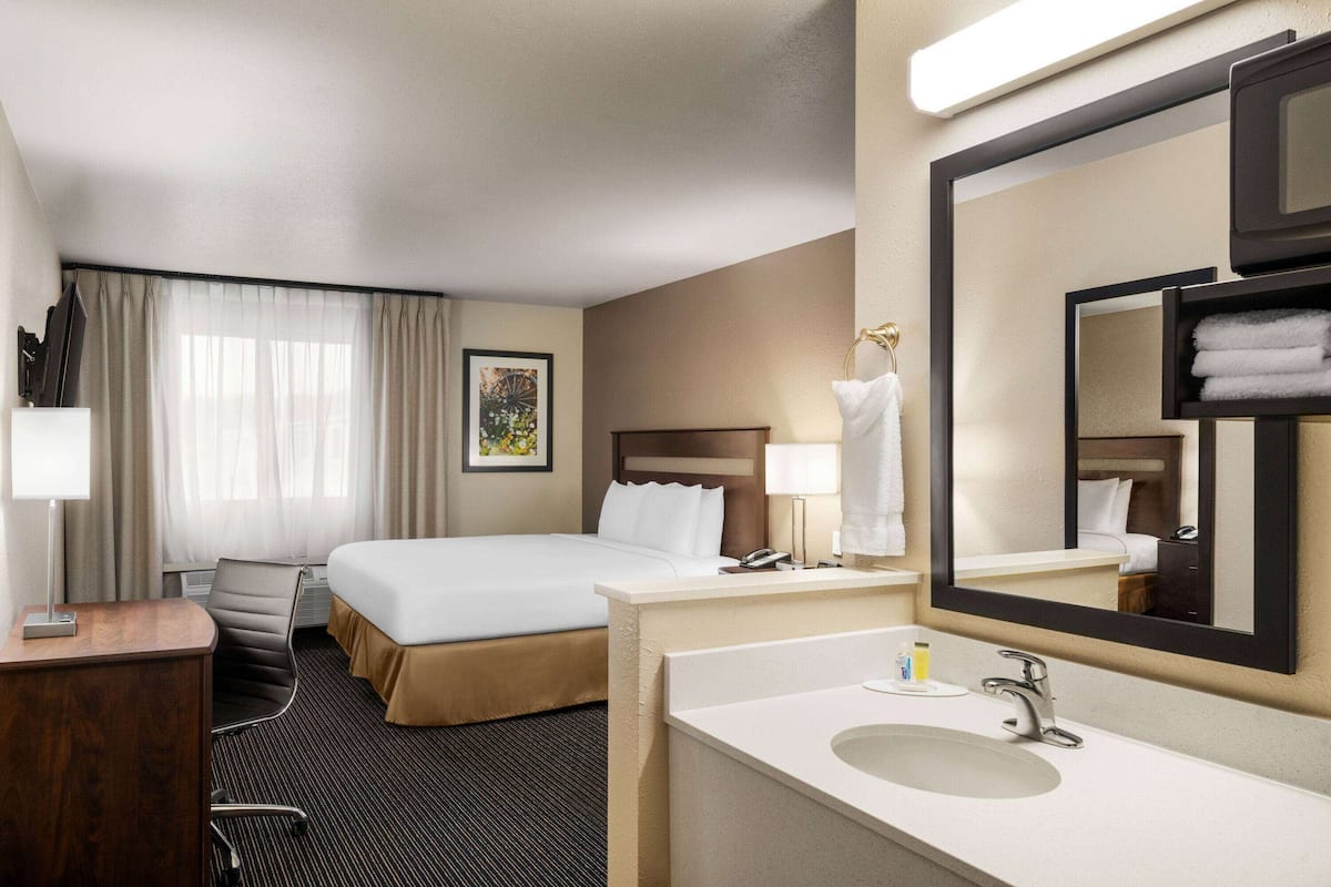 A hotel room with a bed, a desk, a chair, a bathroom with a sink and mirror, and a window with curtains.