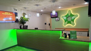 A hotel reception area with a green-lit reception desk, a clock, a security camera, a television, and a sign indicating complimentary breakfast hours. - Extended Stay America Suites Houston Med Ctr NRG Park Kirby (Houston)