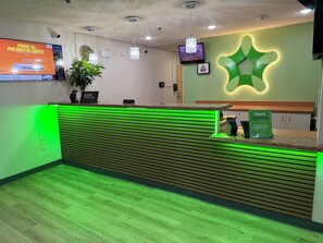 Un hall d’hôtel avec un comptoir d’accueil éclairé en vert, une horloge, une caméra de surveillance, un téléviseur et un panneau indiquant les heures du petit-déjeuner gratuit.
