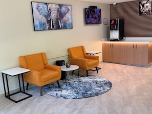 Een moderne zithoek met twee oranje fauteuils, een kleine tafel, een koffieapparaat en een groot abstract schilderij aan de muur.