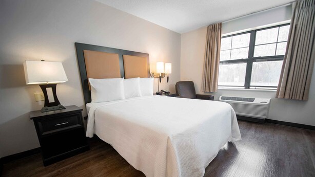 Une chambre d’hôtel avec un grand lit, une table de chevet avec une lampe, une chaise, une fenêtre avec des rideaux et un climatiseur fixé au mur.
