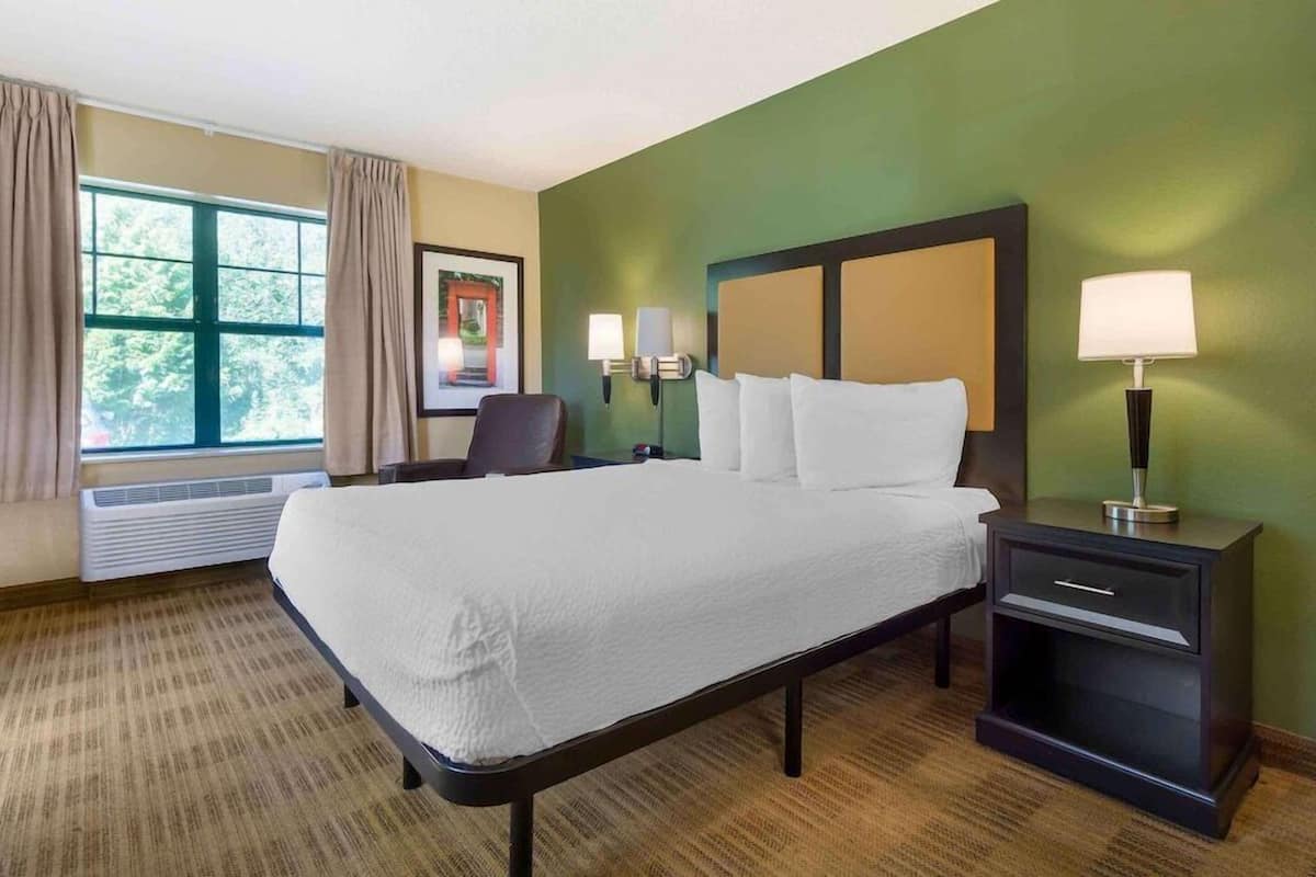 A hotel room with a bed, bedside lamps, a chair, a window with curtains, and a framed picture on the wall.