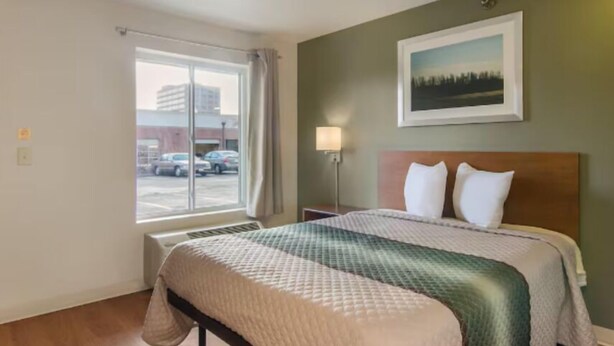 A hotel room with a bed, a window with a view of parked cars, a wall-mounted picture, and a nightstand with a lamp. - Birch Hill Suites Sulphur (Sulphur)