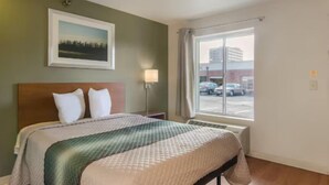 A hotel room with a bed, a window with a view of parked cars, a wall-mounted picture, and a nightstand with a lamp. - Birch Hill Suites Sulphur (Sulphur)