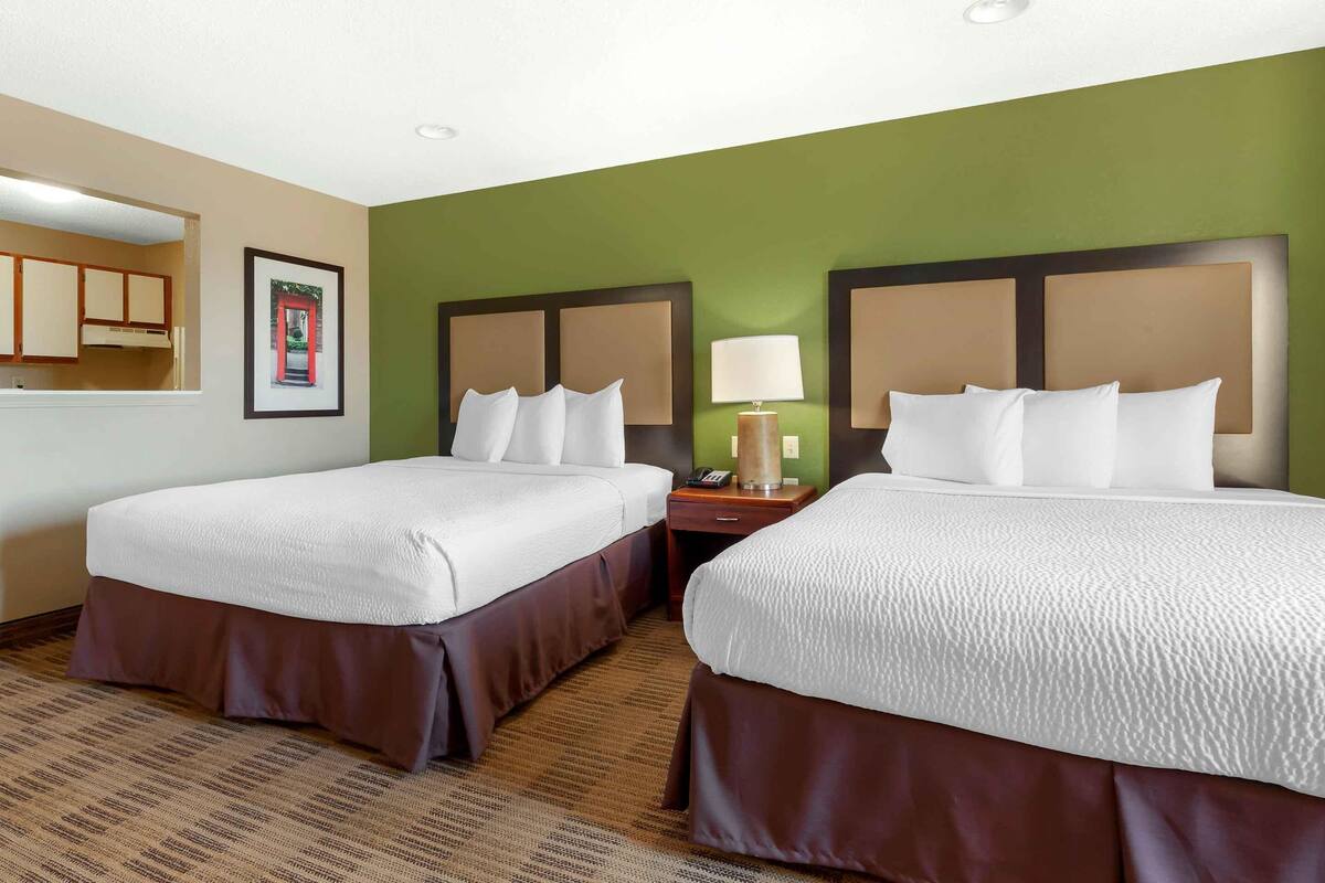 A hotel room with two beds, a nightstand with a lamp, a framed picture on the wall, and a kitchen area visible through an open door.