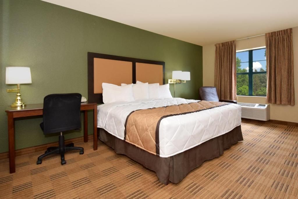A hotel room with a bed, desk, chair, lamps, and a window with curtains.